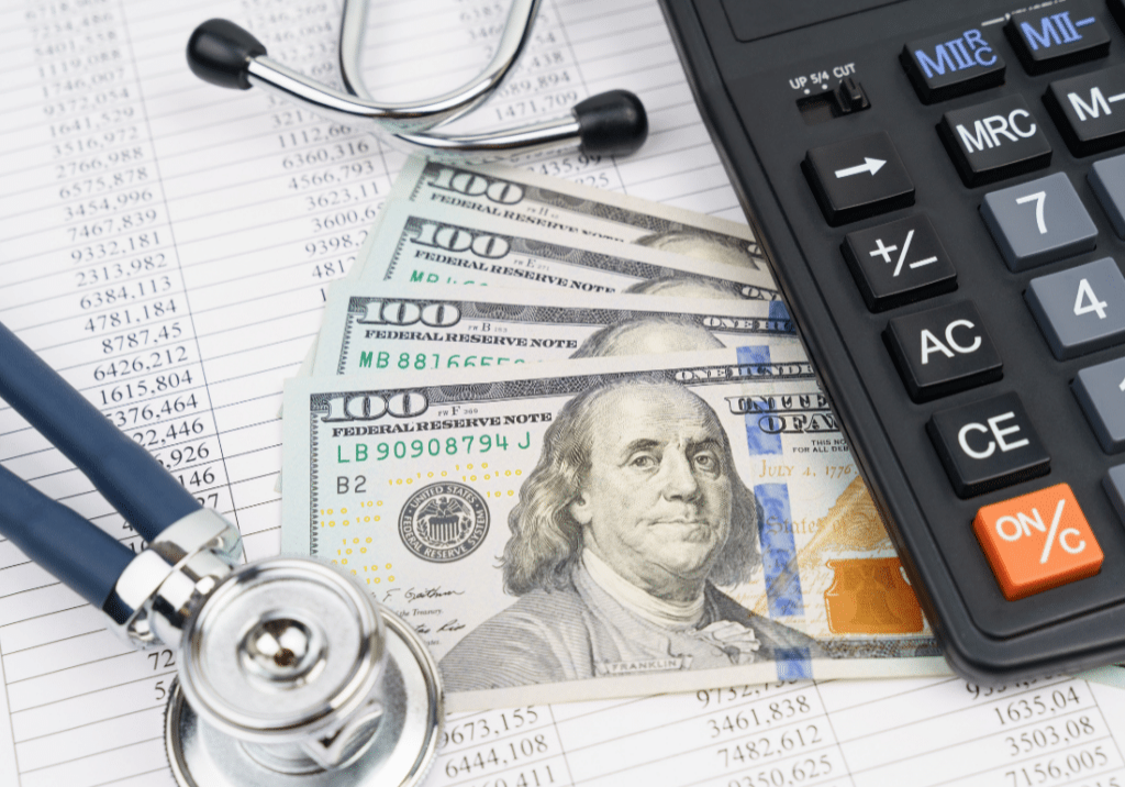 Fighting Inflation featured image 1100 x 800 - 2023-03-24T121053.361 A stethoscope, a calculator, and cash in the form of one hundred dollar bills are pictured on top of financial documents.