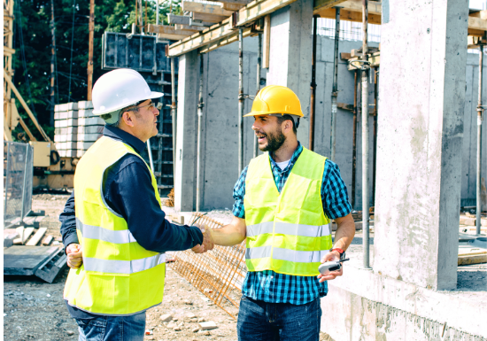 Two construction workers shake hands at a construction site. They are standing outside a partially constructed building with construction equipment around them.
