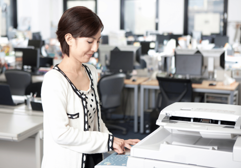 A woman is using a copy machine in an office setting. She is facing the copier while smiling and pressing a button.