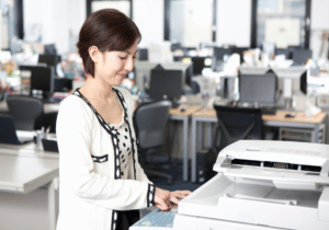 GASB 87 implementation featured img 1100 x 800 A woman is using a copy machine in an office setting. She is facing the copier while smiling and pressing a button.