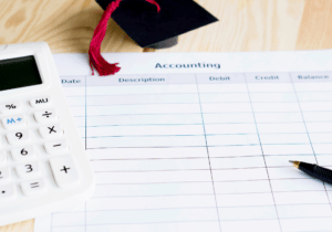 A calculator, pen, and small graduation cap are on top of piece of paper that appears to be a financial statement.