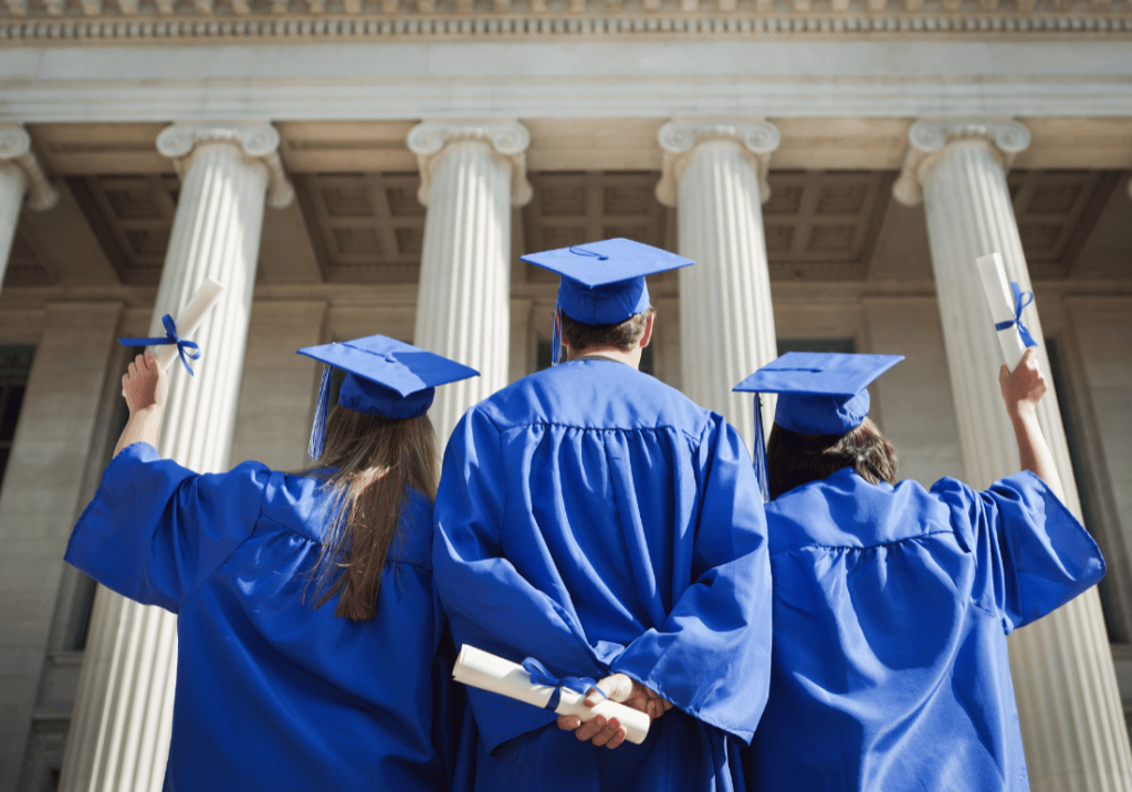 Higher Education Under the IRA featured image Three college graduates, wearing a cap and gown, stand outside a building on a university campus.