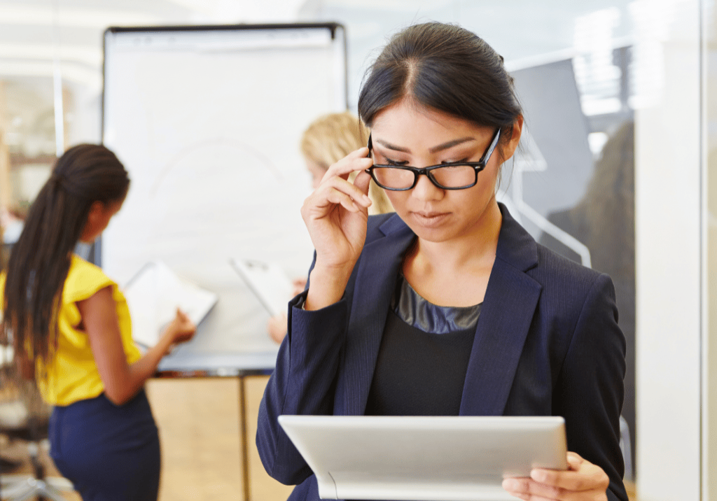 A business woman wearing a suit and glasses is reading a sheet of paper while two others our assessing a white board in the back ground.
