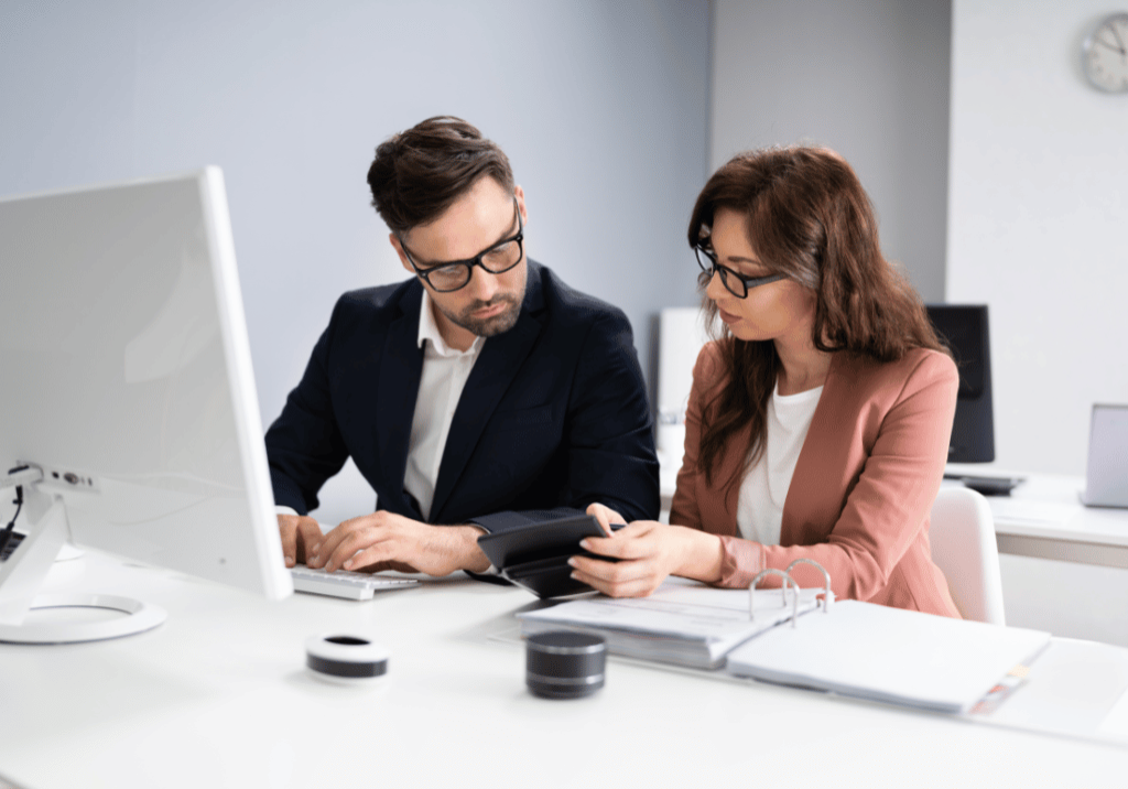 Two accountants at a desk with a computer and an open binder. Both of them are looking at a calculator while one of them is typing.