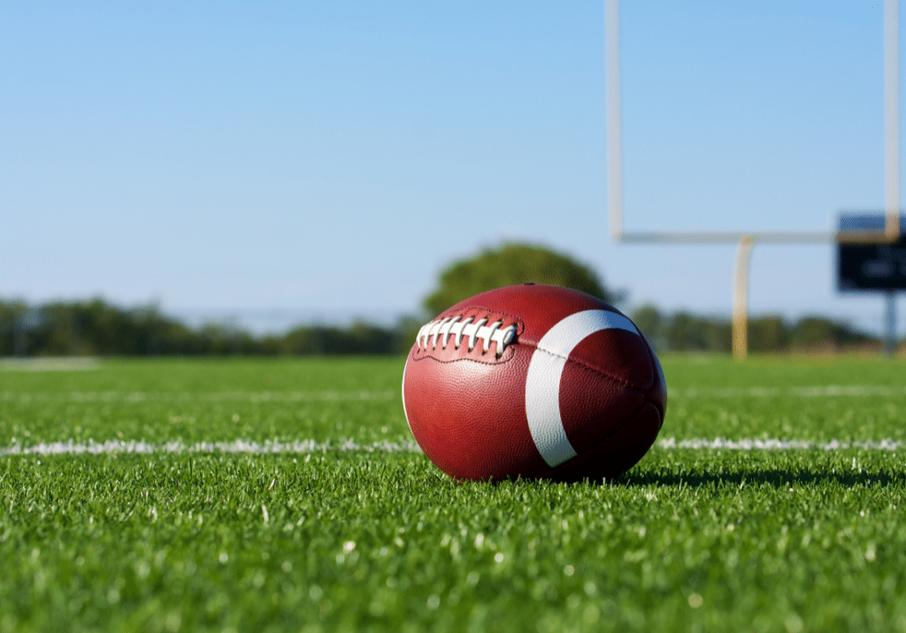 A football is shown close up on football field. The goalposts our visible in the background.