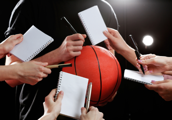 An athlete signs a basketball while fans hold out pens and pads of paper, waiting for their turn to get an autograph.
