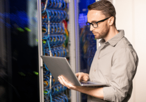 An IT professional is standing in a server room evaluating the IT servers with his laptop.