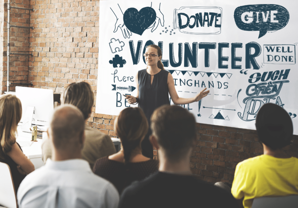 Purpose-Driven Culture featured image 1100 x 800 (90) A person is speaking in front of a group of people. She is standing at the front of a crowd and pointing toward a banner on the wall that says "Volunteer."