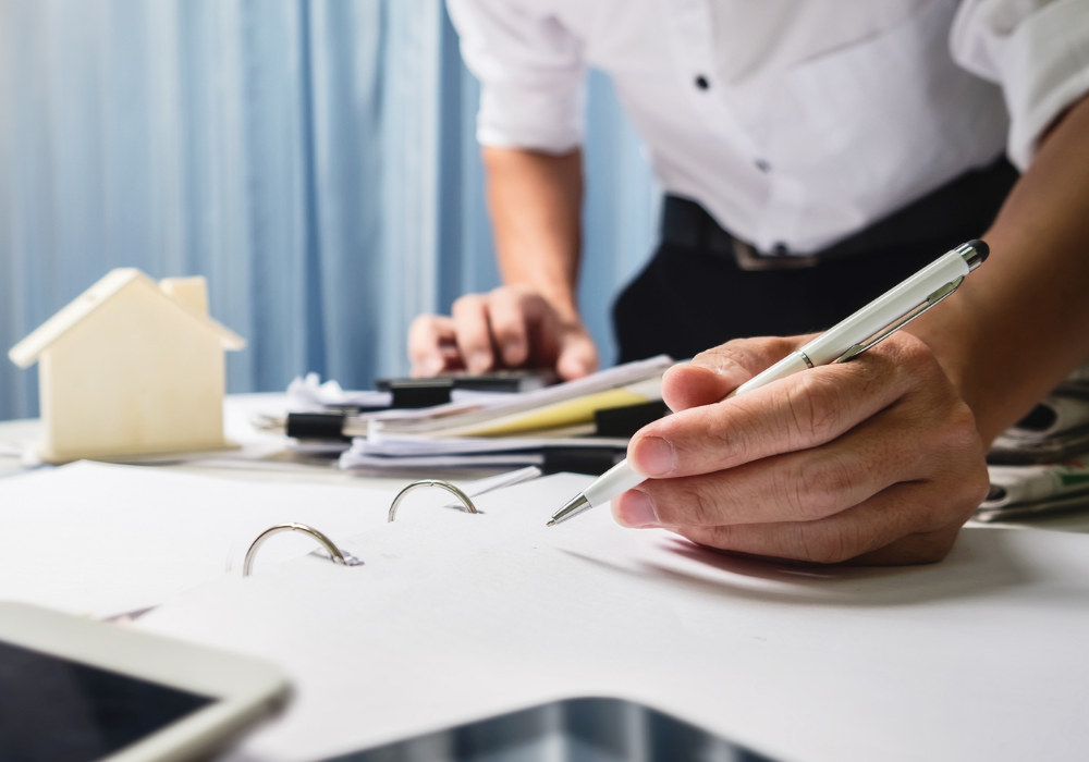 Real Estate bookkeeping_eblast img (1) A man is standing over a table and writing in an open book. On the table is a small model of a house, a calculator, and a tablet device. The man has a pen in his hand and is typing on the calculator with his other hand.