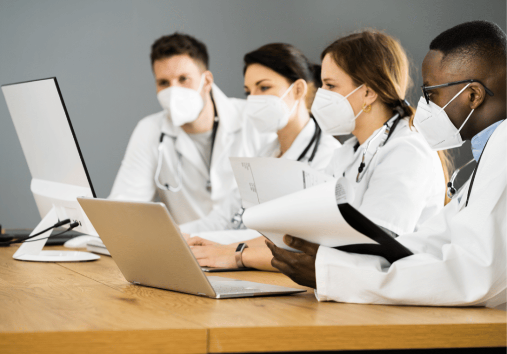 Stark Law Regulations_featured image 1100 x 800 Four physicians are sitting at a table looking at a computer screen and a laptop. All four physicians are wearing a mask and one is holding a clipboard and papers..