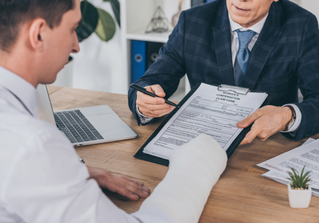 The 411 on Workers' Comp 1100 x 800 An insurance agent is handing a worker's compensation claim form to an injured worker. The injured worker is sitting at a desk with a cast on his arm and readfing the form. The insurance agent is sitting accros the desk, pointing at the form with his pen and explaining what it says.