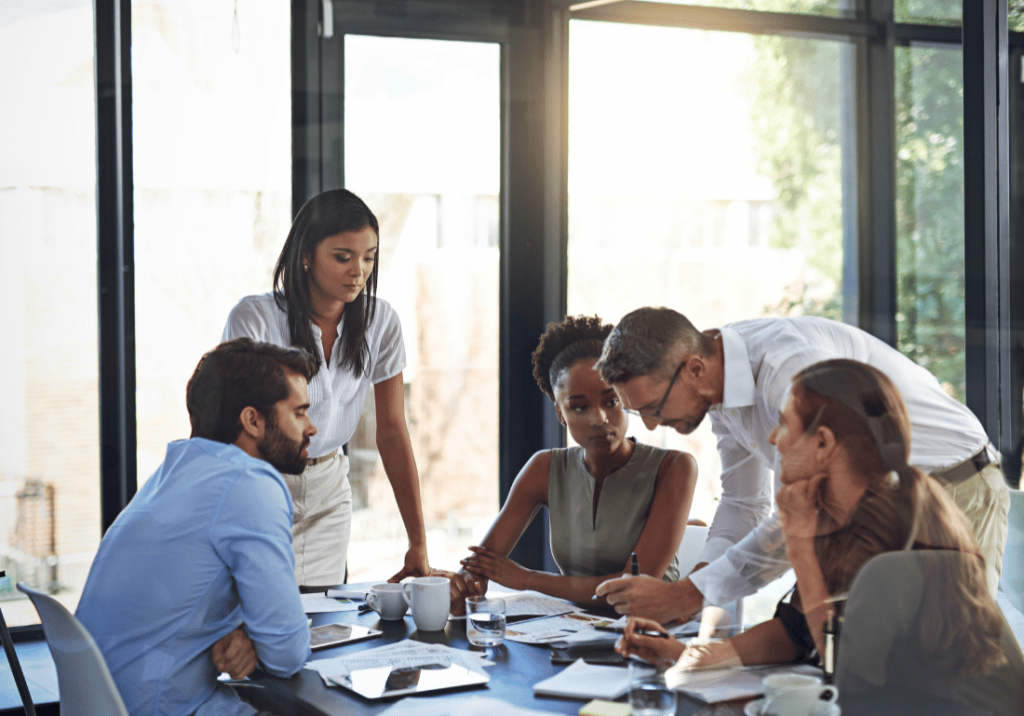 The Basics of Revenue Recognition featured image 1100 x 800 (33) 5 people are gathered around a table ion an office building. One of them is writing on paper while the others look on. There are papers and coffee mugs on the table and a large window in the background.