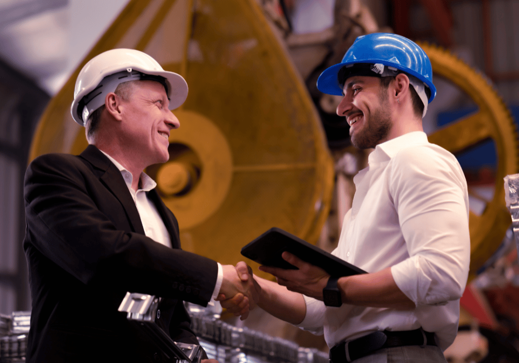 a business owner and employee smile and shake hands in a manufacturing facility. Both are wearing hard hats and there is machine equipment in the background.