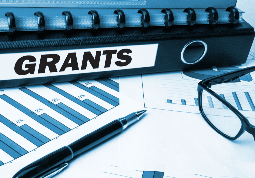 A binder that says "Grants", a pen, and glasses shown on a background of documents. The documents show charts and numbers.