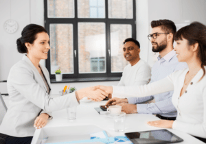 A business person is sitting across a from three employees at a table in an office setting. The business person is shaking hands with one of the employees.
