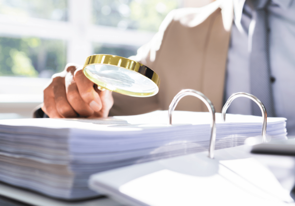 audit red flags featured image 1100 x 800 (64) An auditor reviews documents in a binder with a magnifying glass.