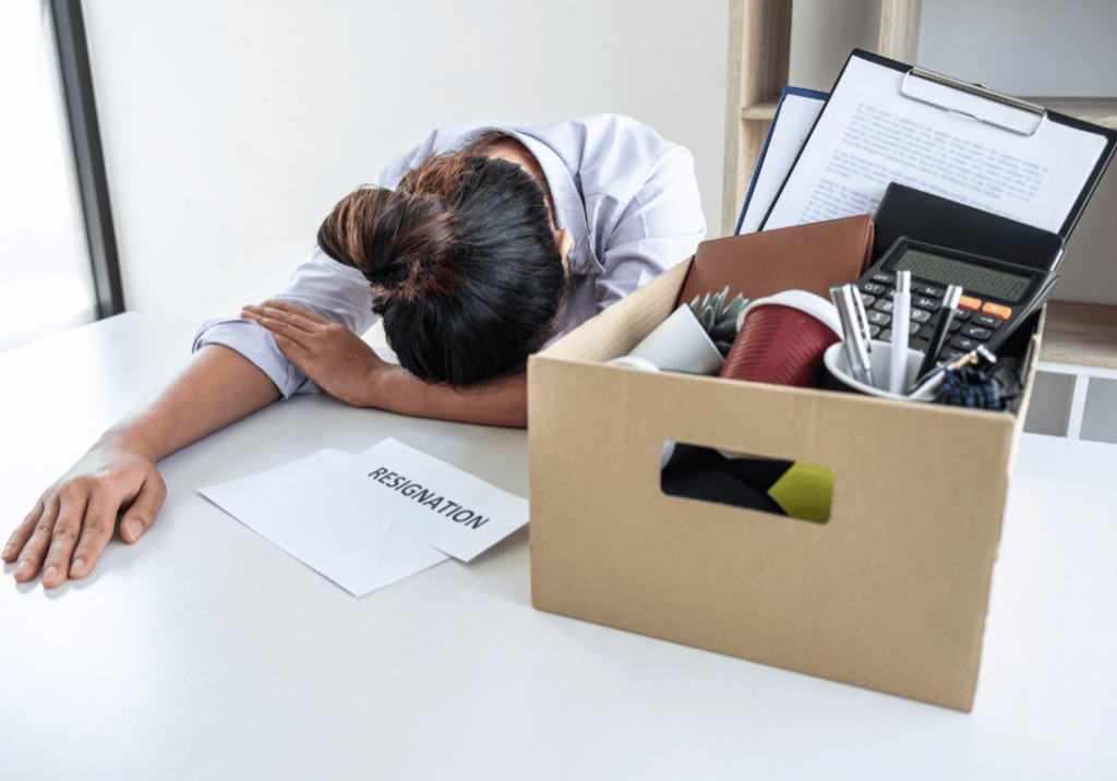 A box packed with office supplies and a resignation letter on a desk next to a disengaged employee who has their head down.