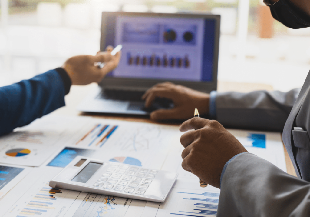 featured image 1100 x 800 - 2023-05-17T123348.533 Two business persons are sitting at a table and pointing toward an open laptop computer. There are documents, graphs, and a calculator on the table. The laptop screen is also showing a graph.