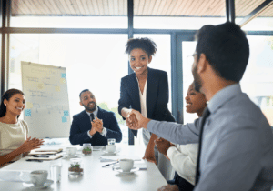 featured image 1100 x 800 (3) People dressed in business attire are in an office room sitting at a table. Two of them are shaking hands and the rest are applauding.