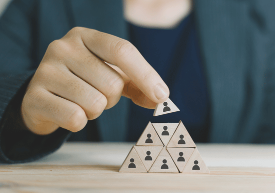 featured image (18) The image shows someone wearing a business suit stacking triangle shaped wood pieces into a pyramid shape. The triangle pieces show a stick figure person on each piece. The business person's face is not shown and the image is focused on their hand and the triangle pieces.