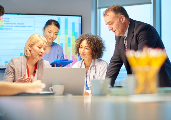 Two peole are sitting at a table looking at a laptop computer. A man and a woman are standing behind them looking at the same screen. There is a screen in the background that is displaying a chart. Coffee mugs and pencils are on the table in front of the people.