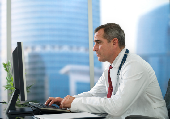 A physician sits at his desk looking at a computer and appears focused on the computer screen. Highrise buildings are visible through his office window.